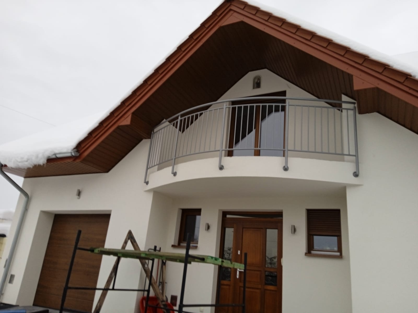 Nowoczesny dom jednorodzinny z balkonem zabezpieczonym metalową balustradą w kolorze szarym, widoczne rusztowanie przed wejściem.