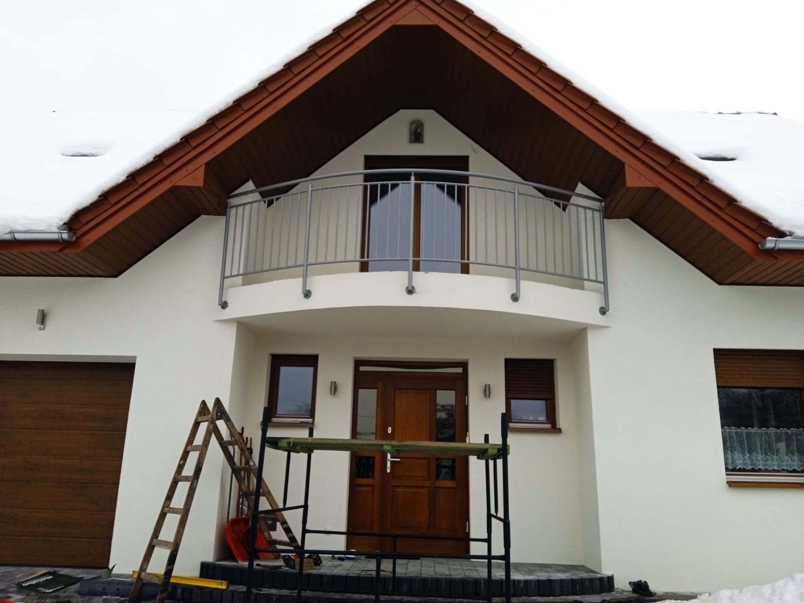 Dom jednorodzinny z balkonem zabezpieczonym szarą, metalową balustradą, widoczne rusztowanie i drabina wskazujące na prace wykończeniowe.