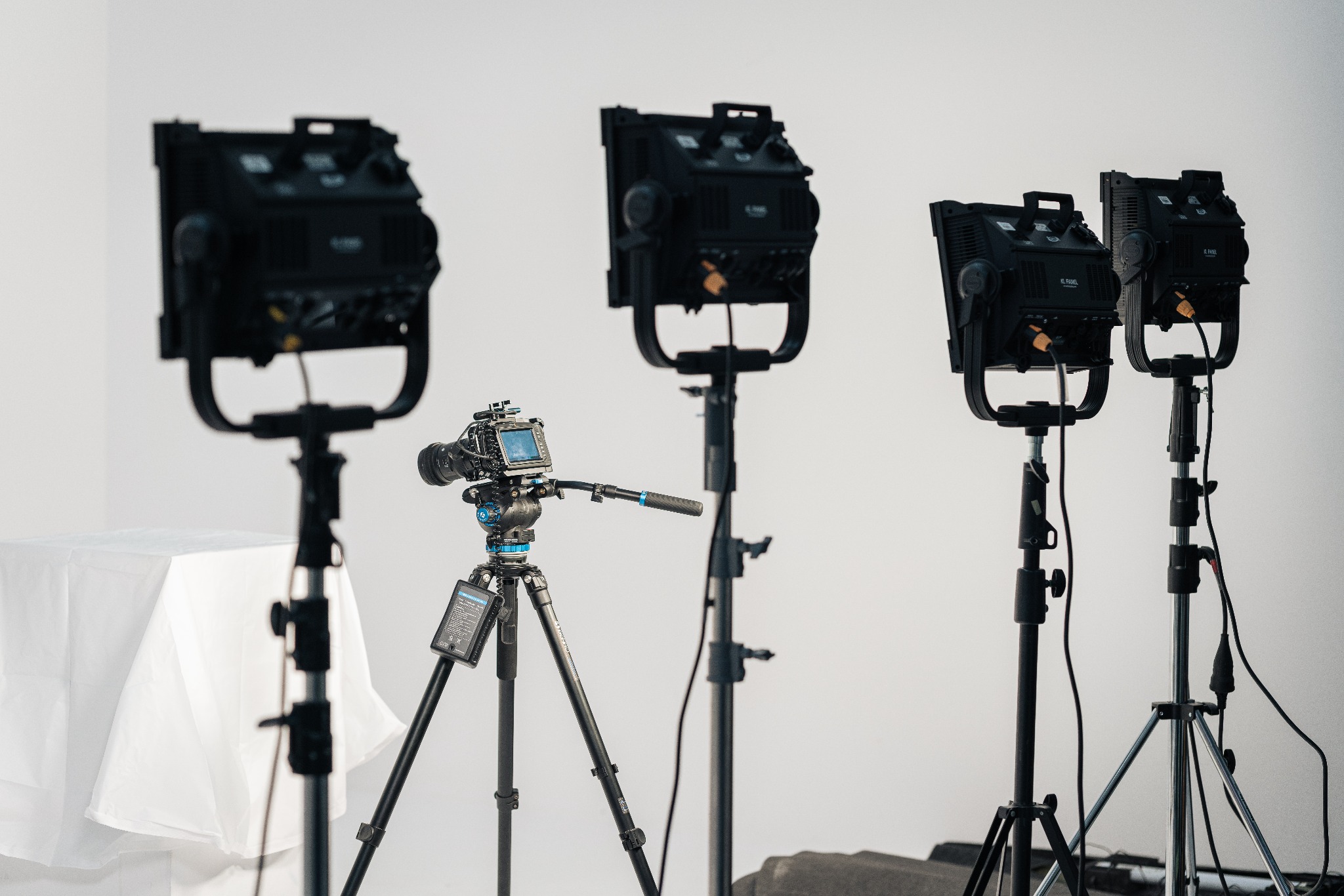 Profesjonalny sprzęt filmowy: kamera na statywie i trzy lampy studyjne na statywach przed białym tłem, przygotowane do filmowania.
