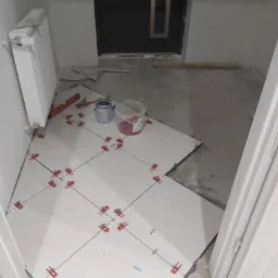 Układanie jasnych płytek podłogowych w przedpokoju, widoczne krzyżyki dystansowe, poziomica i pojemniki z zaprawą, częściowo zakończone prace.