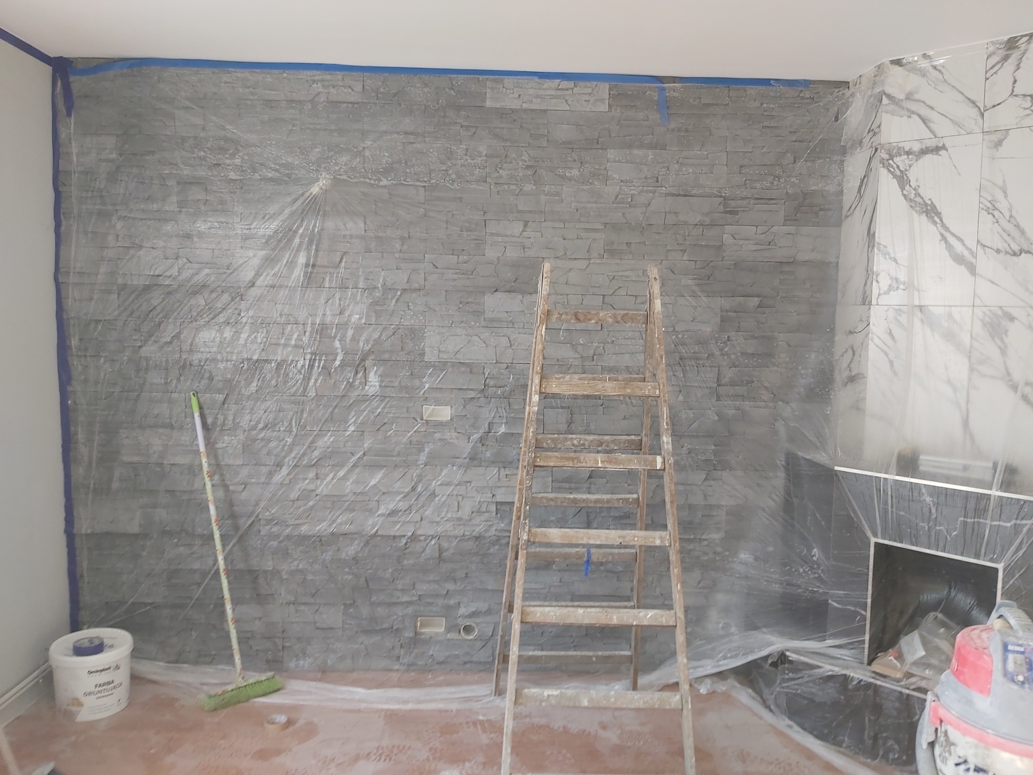 Montaż kamienia dekoracyjnego na ścianie w Jarocinie, zabezpieczony folią malarską, z widoczną drabiną, wiadrem z farbą gruntującą i odkurzaczem przemysłowym.