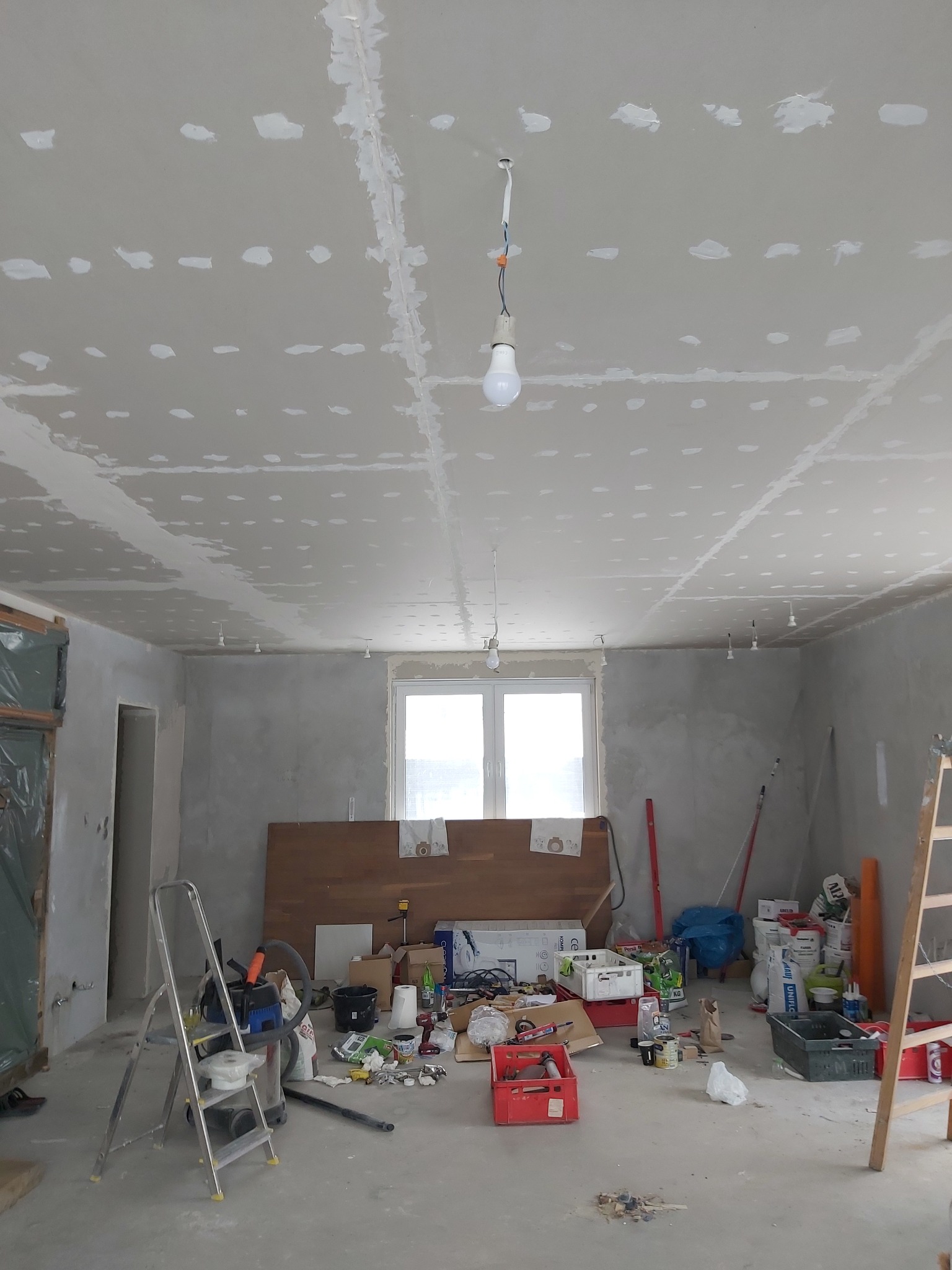 Wnętrze w trakcie remontu, sufit i ściany z płyt kartonowo-gipsowych, widoczne łączenia płyt wypełnione masą szpachlową, prowizoryczne oświetlenie żarówkami, liczne narzędzia i materiały budowlane...