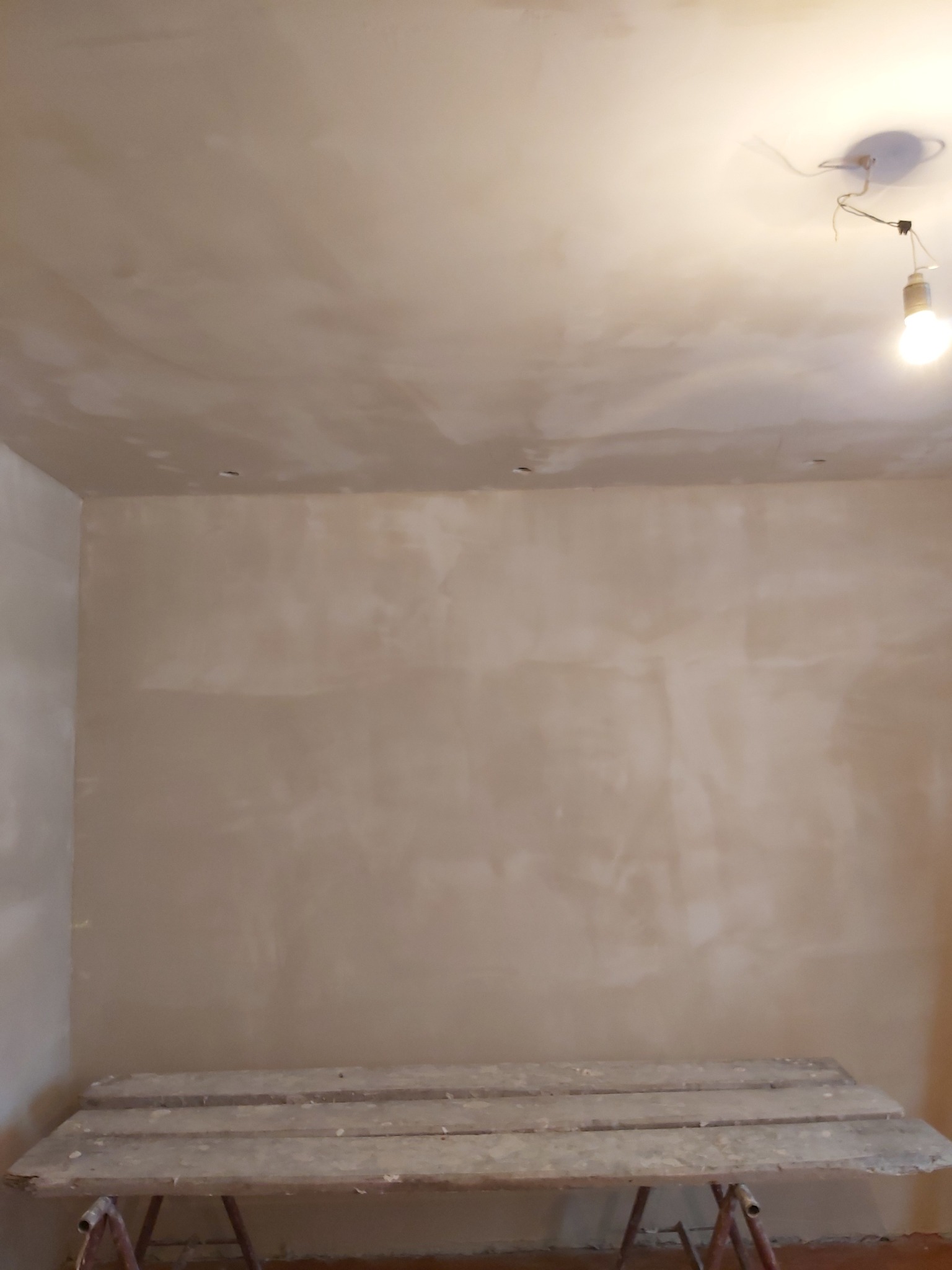 Ściana i sufit po nałożeniu warstwy wygładzającej, widoczny stół roboczy, surowa żarówka i niedoskonałości na powierzchni.
