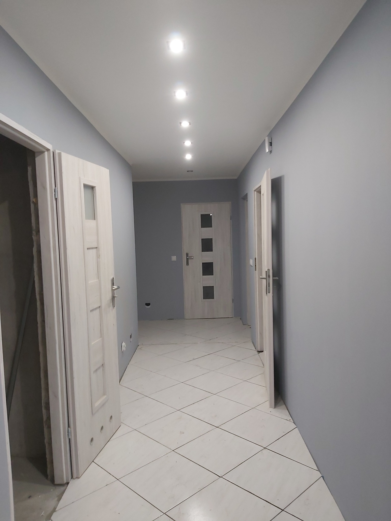 Szary korytarz po remoncie z białymi drzwiami, białymi płytkami podłogowymi ułożonymi w karo i rzędem punktowych lamp sufitowych.