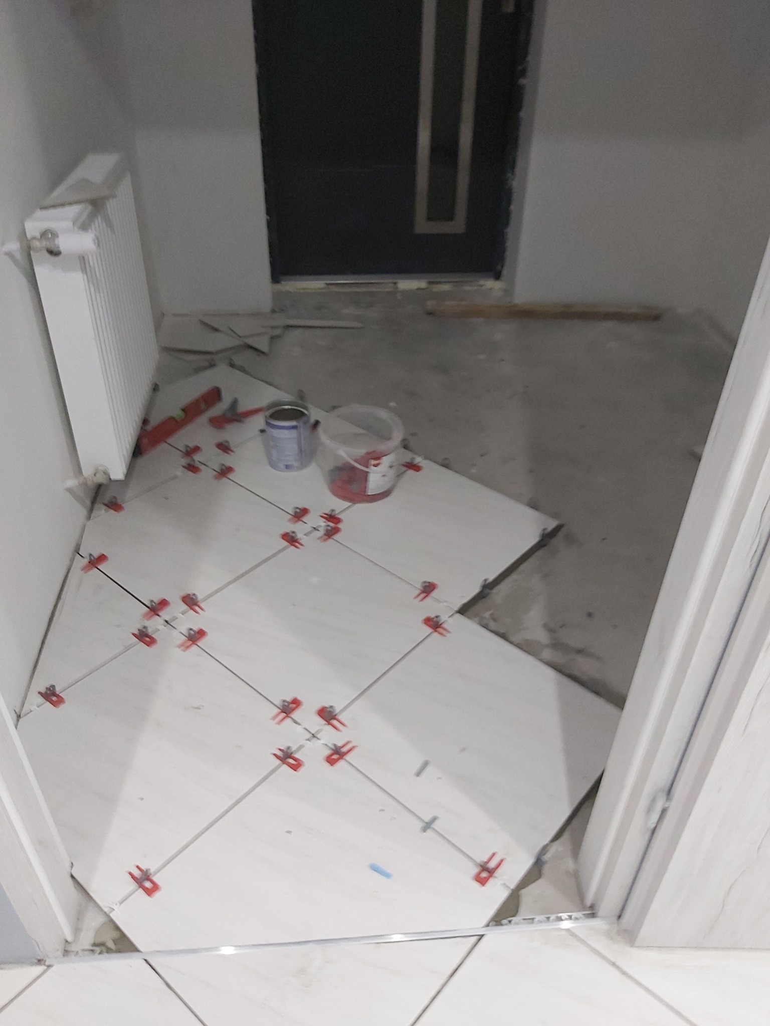 Układanie jasnych płytek podłogowych w przedpokoju, widoczne krzyżyki dystansowe, poziomica i pojemniki z zaprawą, częściowo zakończone prace.