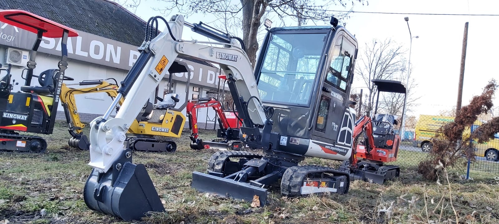 Grupa nowych minikoparek marki Kingway różnych kolorów zaparkowana na placu przed budynkiem salonu sprzedaży.