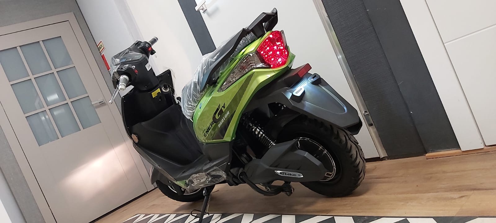 Nowy, zielono-czarny skuter elektryczny Barton Energy, zaparkowany w pomieszczeniu z białą stolarką drzwiową i podłogą w czarno-białą szachownicę, widok z tyłu.