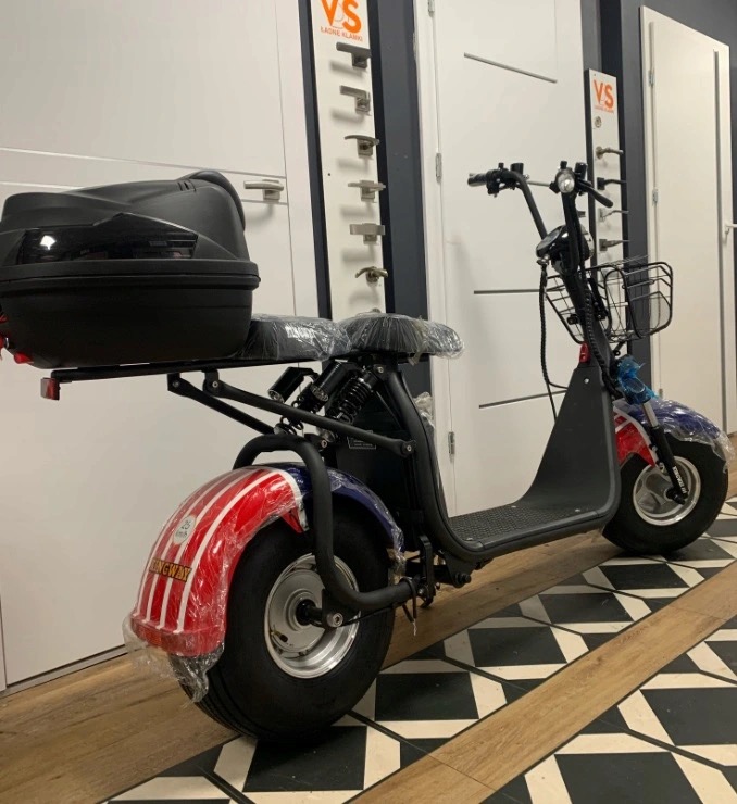 Elektryczny skuter miejski z dwoma siedzeniami i koszykiem z przodu, zaparkowany na podłodze w szachownicę, z tyłu widoczna czarna skrzynia bagażowa i ekspozycja klamek do drzwi.