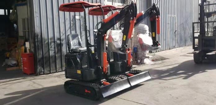 Dwie nowe, czarne minikoparki z pomarańczowymi akcentami, częściowo owinięte folią ochronną, stoją obok siebie na betonowym placu, w tle blaszana hala i wózek widłowy.