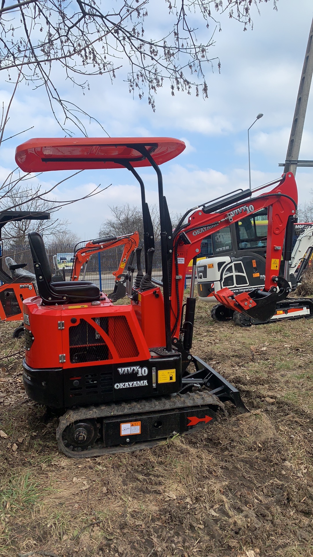 Czerwona minikoparka OKAYAMA VIVY10 na gąsienicach, z podniesionym ramieniem i łyżką, stojąca na ziemi z suchą trawą, inne maszyny w tle.