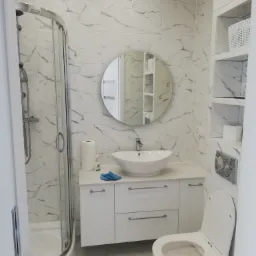 Nowoczesna łazienka z kabiną prysznicową, umywalką nablatową na białej szafce, okrągłym lustrem i toaletą, wykończona płytkami imitującymi marmur z subtelnym wzorem falistym w górnej części ściany.