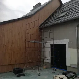 Elewacja domu w trakcie prac wykończeniowych: część ściany pokryta panelami imitującymi drewno, druga część w trakcie ocieplania, rusztowanie przy ścianie, widoczne materiały budowlane.