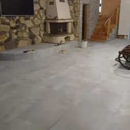 Salon z nową, jasnoszarą podłogą z płytek, kamiennym kominkiem i drewnianymi schodami, z drewnianym fotelem bujanym