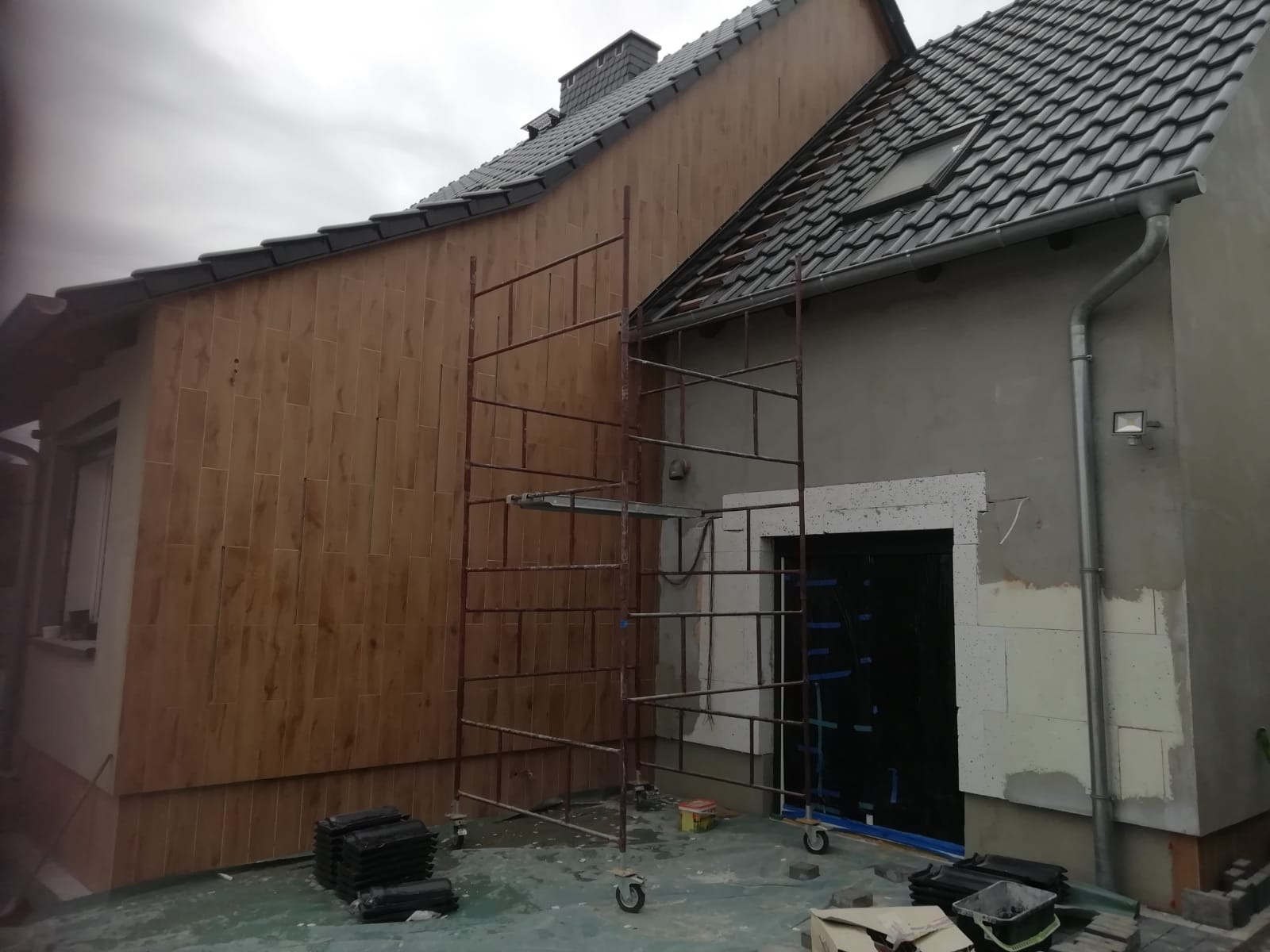 Elewacja domu w trakcie prac wykończeniowych: część ściany pokryta panelami imitującymi drewno, druga część w trakcie ocieplania, rusztowanie przy ścianie, widoczne materiały budowlane.