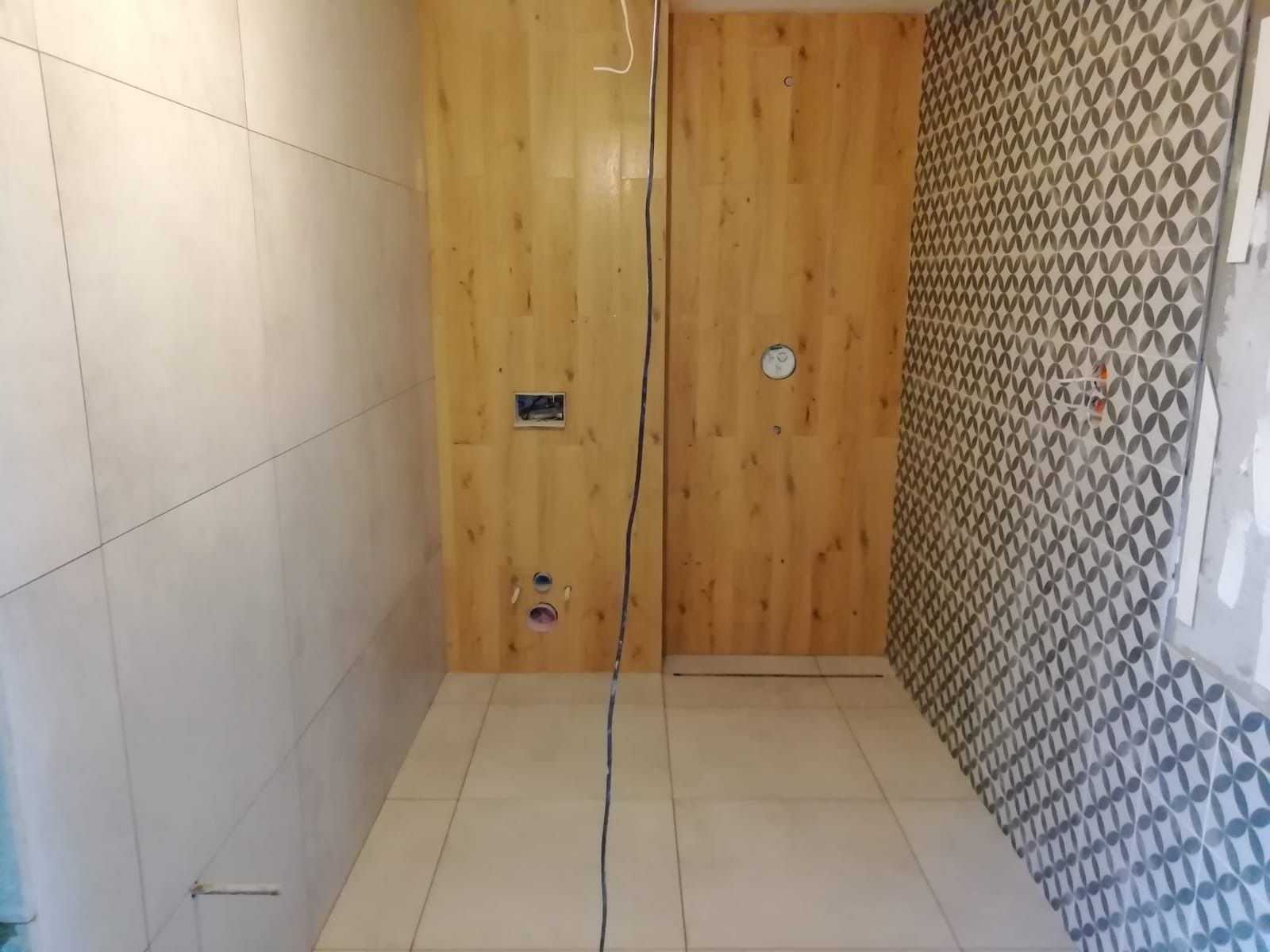 Wnętrze łazienki w trakcie wykańczania: ściany wyłożone beżowymi płytkami, dekoracyjna ściana z wzorem geometrycznym i drewnopodobna okładzina, widoczne otwory pod instalacje i przewody elektryczne.