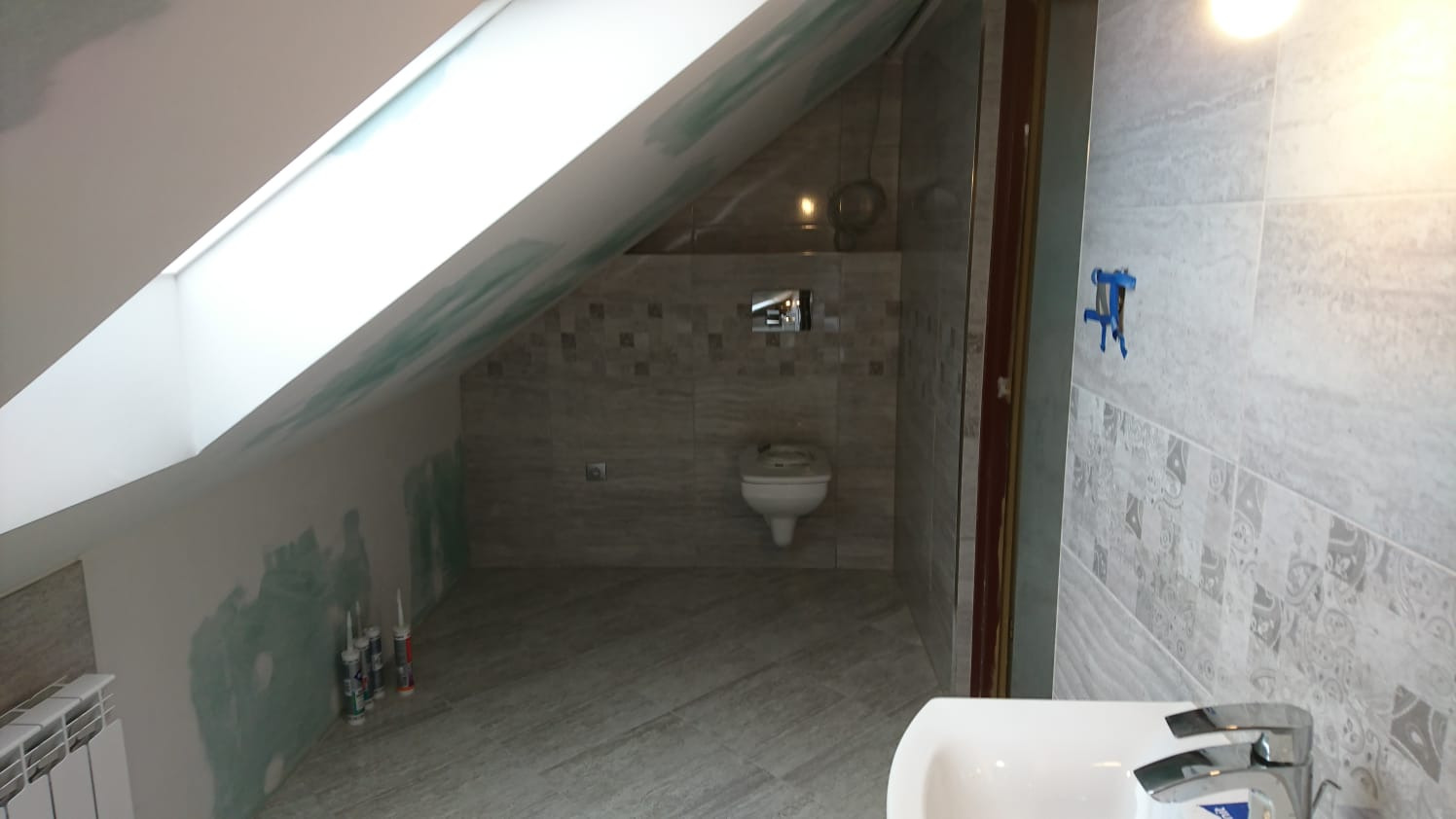 Łazienka na poddaszu w trakcie remontu, widoczne płytki imitujące kamień, częściowo położone, oraz silikonowe tuby na podłodze przy ścianie ze skosem.