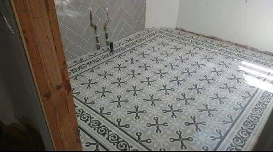 Ukończona podłoga w łazience z dekoracyjnymi płytkami w odcieniach szarości, wzór w kształcie skrzyżowanych kluczy francuskich, ściana wyłożona płytkami w jodełkę, widoczne rury instalacyjne.