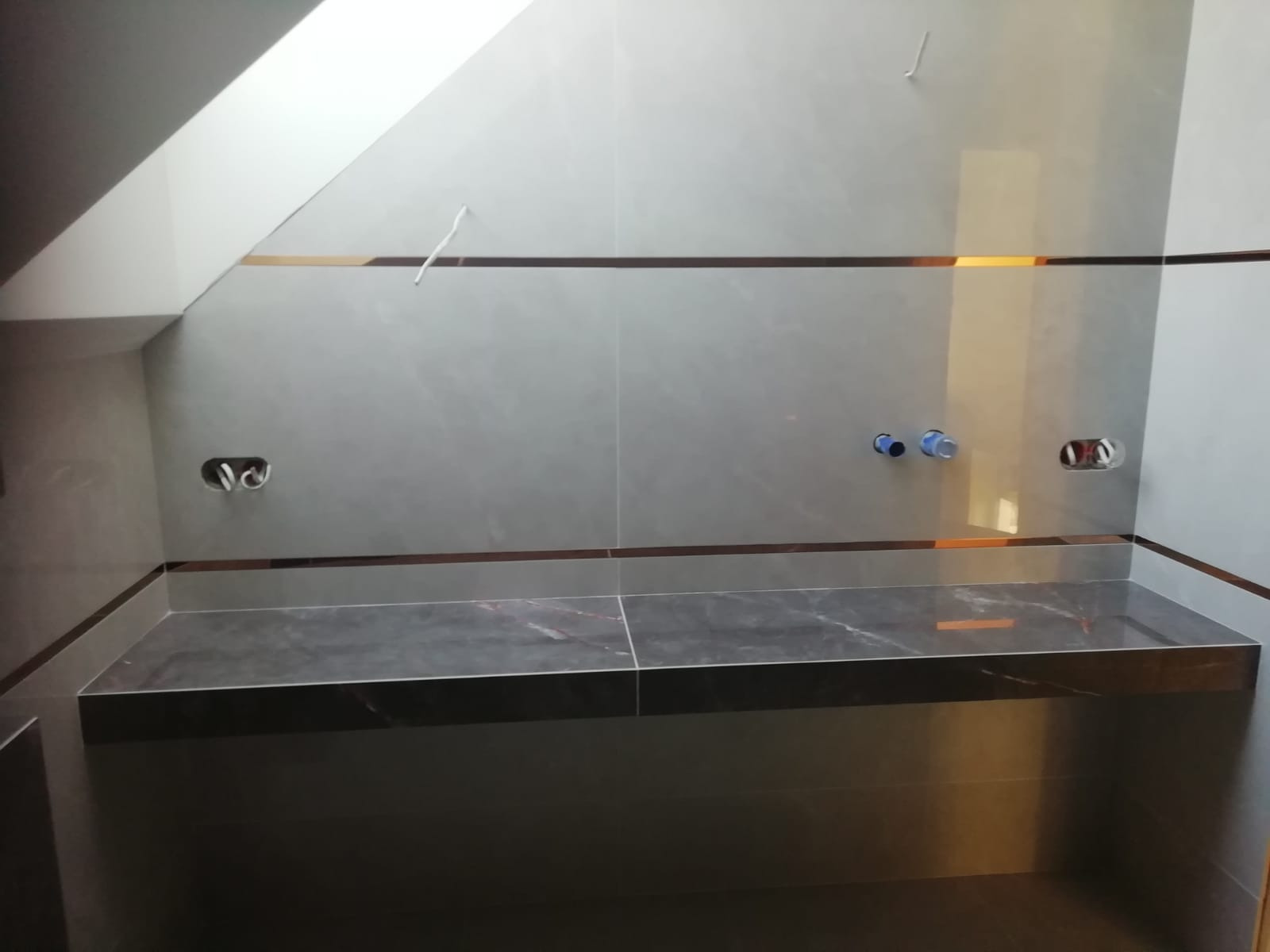 Ściana w łazience w trakcie wykańczania, szare płytki z dekoracyjnymi, poziomymi paskami, przygotowane otwory na armaturę, ciemny blat pod umywalkę.