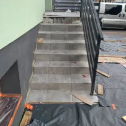 Schody wejściowe do budynku w trakcie układania płytek, zabezpieczone folią ochronną, widoczne narzędzia i resztki materiałów budowlanych.