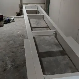 Metalowa konstrukcja ramy, prawdopodobnie pod schody, w surowym stanie, przytwierdzona do ściany, widoczne ślady prac budowlanych i niedoskonałości w wykończeniu.