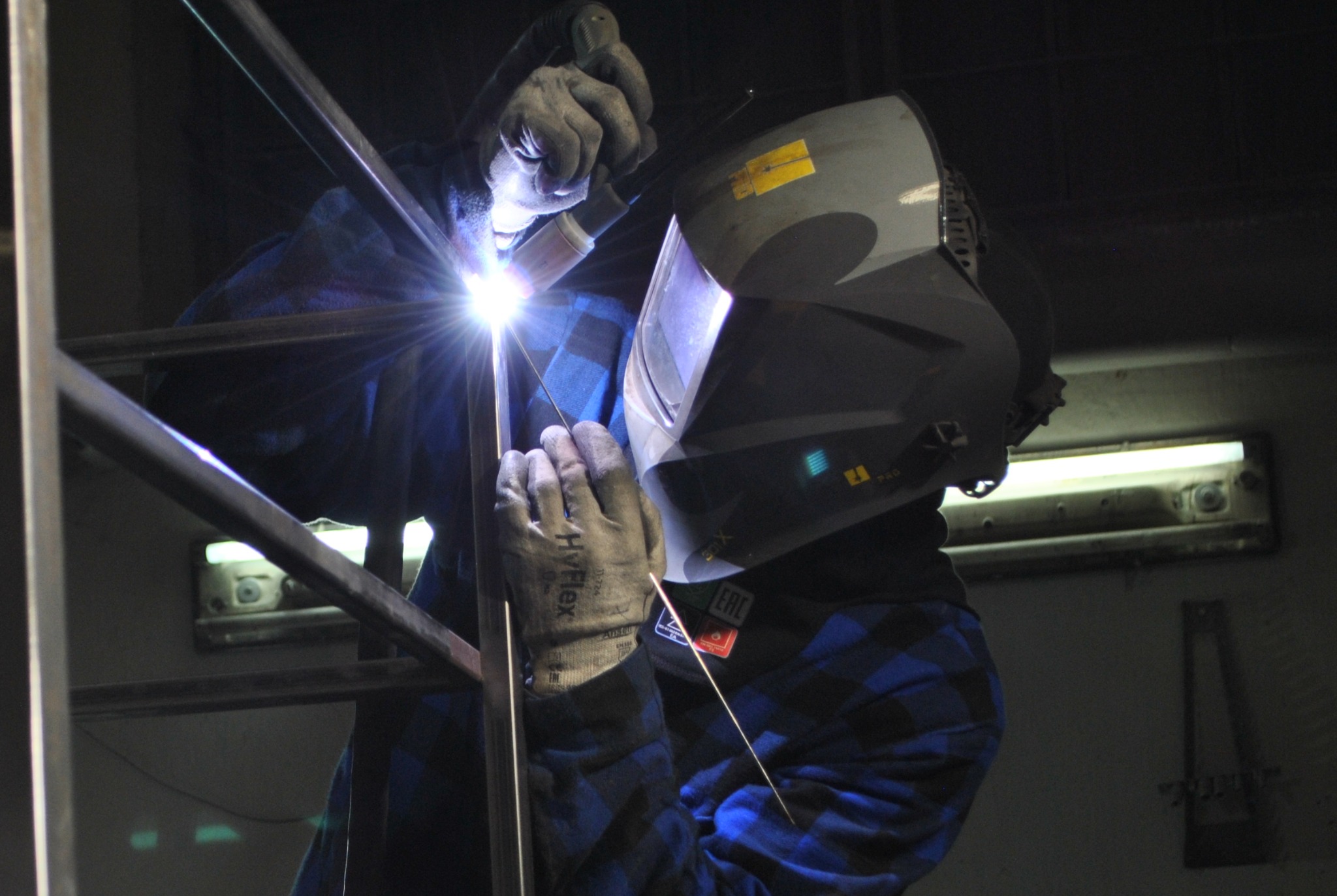Pracownik w masce ochronnej spawa metalową konstrukcję w warsztacie; widoczny jasny błysk spawania i iskry.