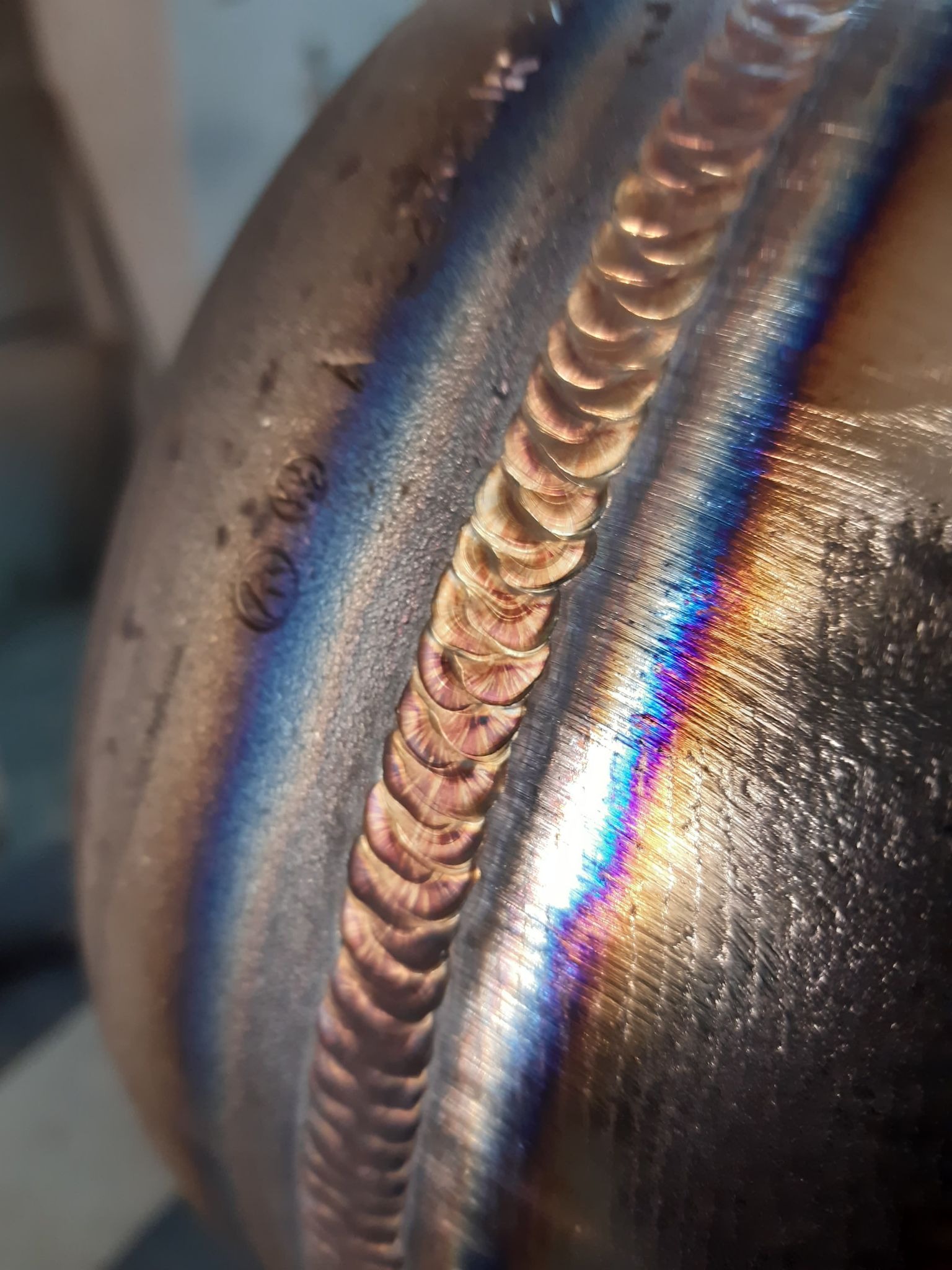 Zbliżenie na świeżo wykonany spaw na metalowej rurze, widoczne charakterystyczne zabarwienia termiczne i drobne nierówności powierzchni.