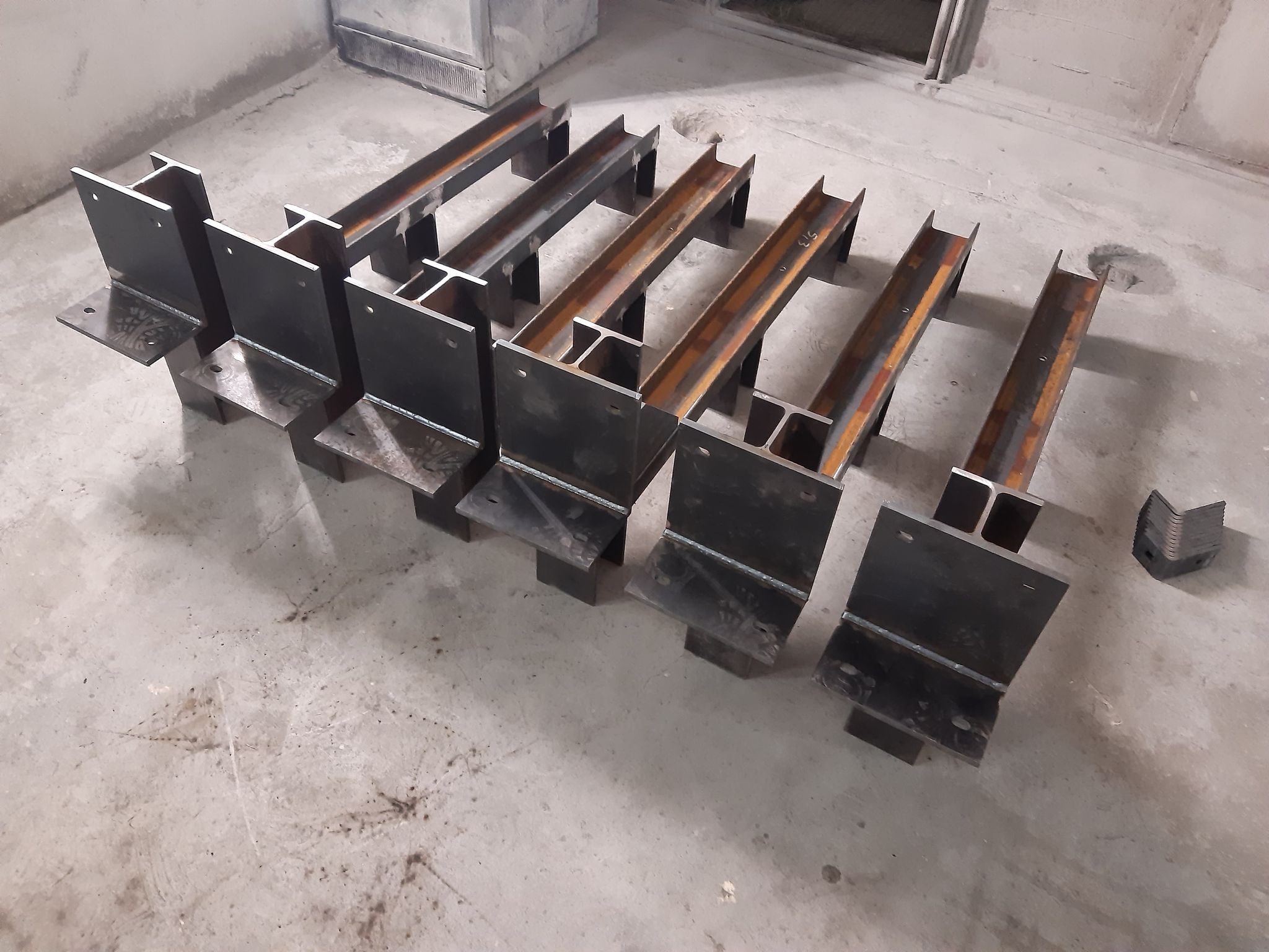 Przygotowane do montażu stalowe elementy konstrukcyjne w kształcie litery H z przyspawanymi podstawami, leżące na betonowej podłodze w surowym wnętrzu.