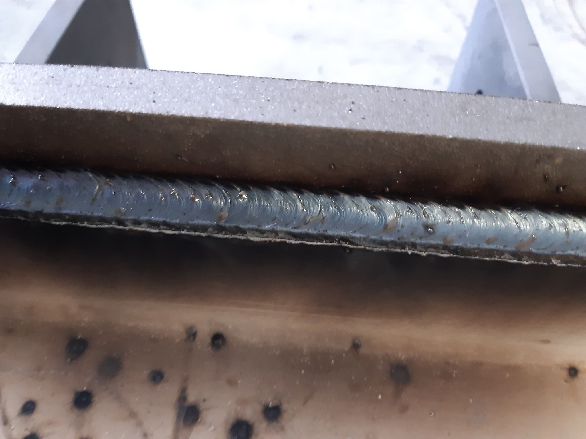 Szczegółowe ujęcie spoiny łączącej dwa elementy metalowe, widoczne nierówności i charakterystyczny wzór po spawaniu, metalowy element w tle.