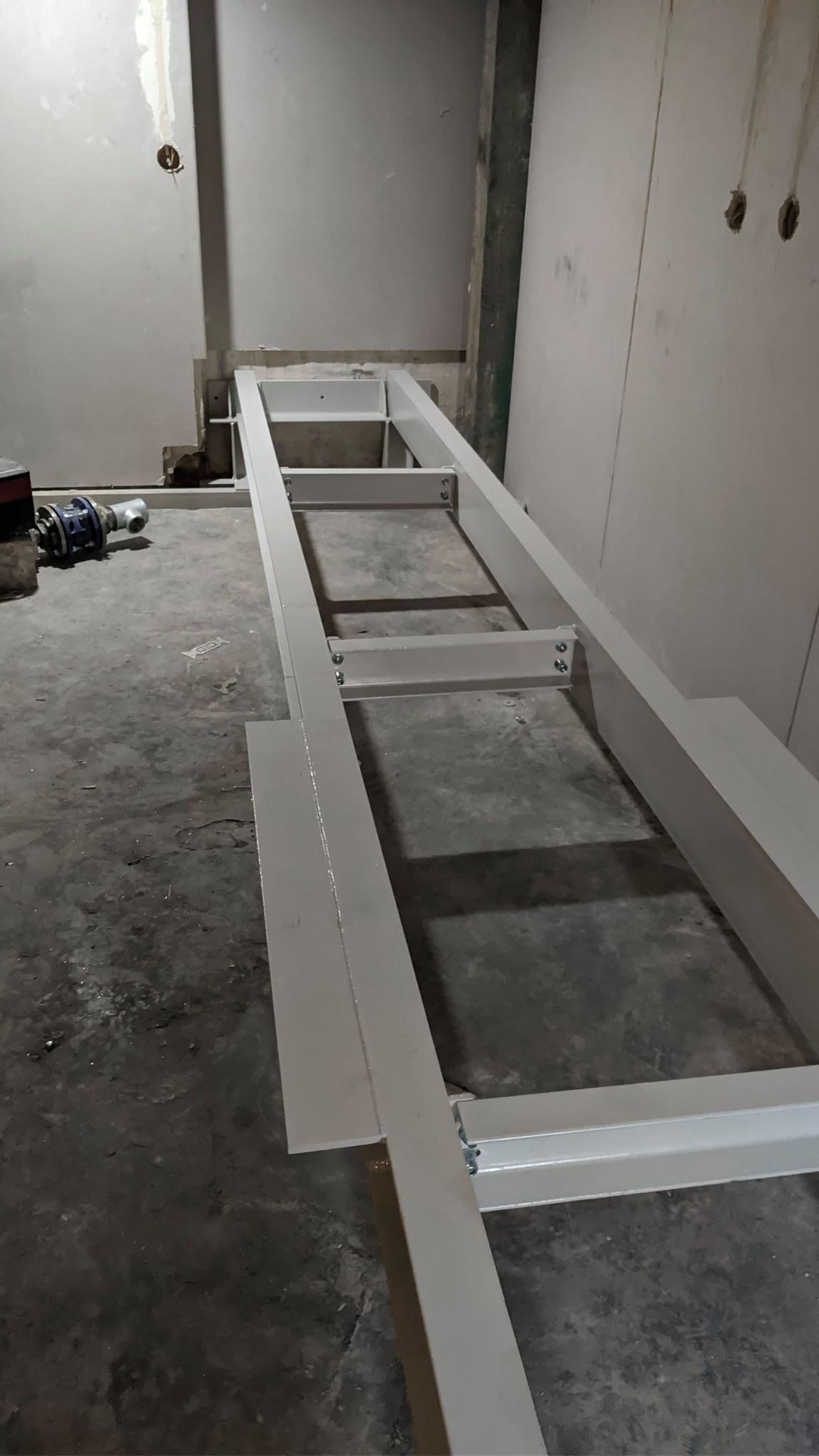 Metalowa konstrukcja ramy, prawdopodobnie pod schody, w surowym stanie, przytwierdzona do ściany, widoczne ślady prac budowlanych i niedoskonałości w wykończeniu.