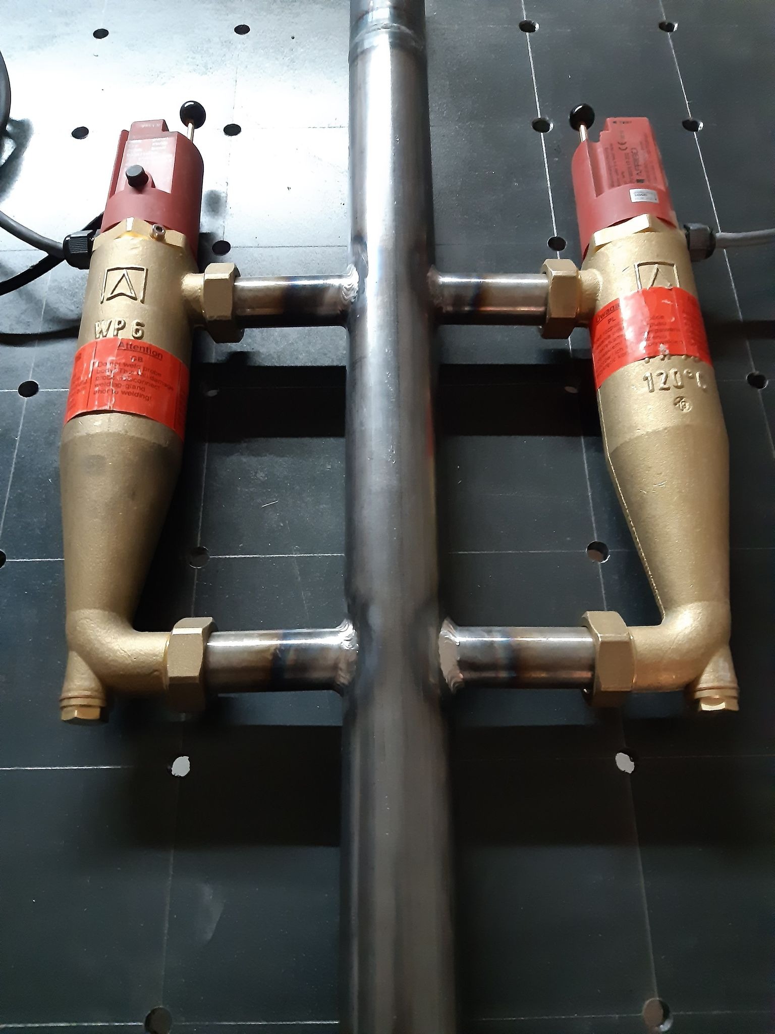 Zespawana stalowa konstrukcja rurowa z dwoma mosiężnymi zaworami termostatycznymi WP6 z czerwonymi etykietami, ustawiona na czarnej płycie montażowej z otworami.