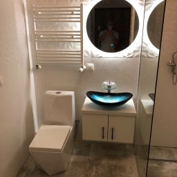 Marek Golebiewski - Nowoczesna łazienka z umywalką w kształcie łodzi w odcieniach błękitu, podświetlanym okrągłym lustrem, grzejnikiem drabinkowym i geometrycznymi płytkami 3D na ścianie. W lustrze odbija się osoba...