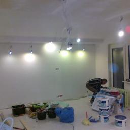 Marek Golebiewski - Pokój w trakcie przygotowań do malowania: widoczne puszki z farbą, narzędzia malarskie i instalacja oświetleniowa na suficie z wiszącymi kablami, mężczyzna pochylony nad pracami przygotowawczymi.