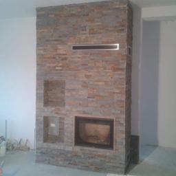 Marek Golebiewski - Obudowa kominka z kamienia dekoracyjnego w trakcie montażu, widoczne otwory wentylacyjne i puste wnęki, w tle ściany w trakcie malowania i narzędzia pracy.