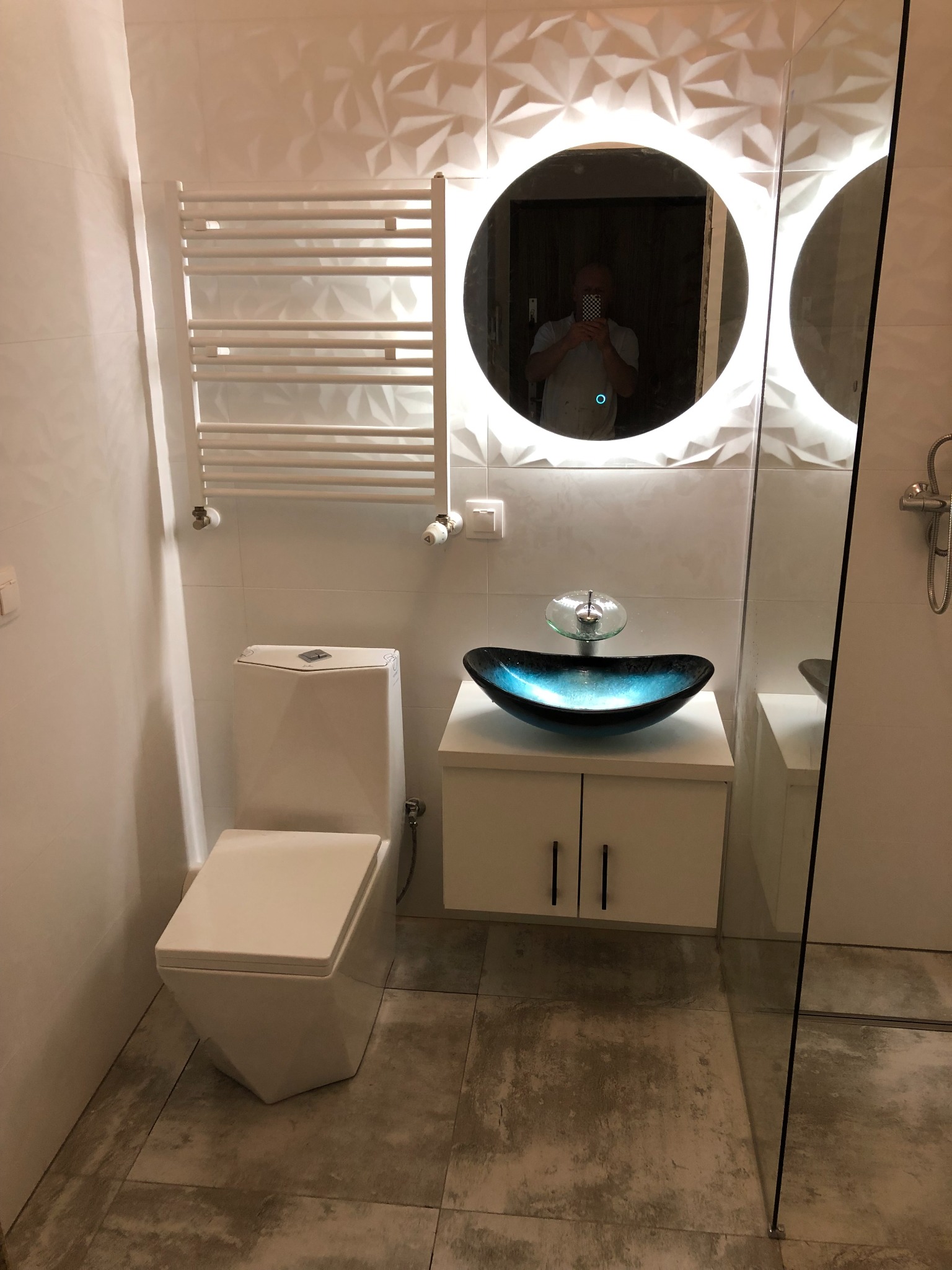 Nowoczesna łazienka z umywalką w kształcie łodzi w odcieniach błękitu, podświetlanym okrągłym lustrem, grzejnikiem drabinkowym i geometrycznymi płytkami 3D na ścianie. W lustrze odbija się osoba...