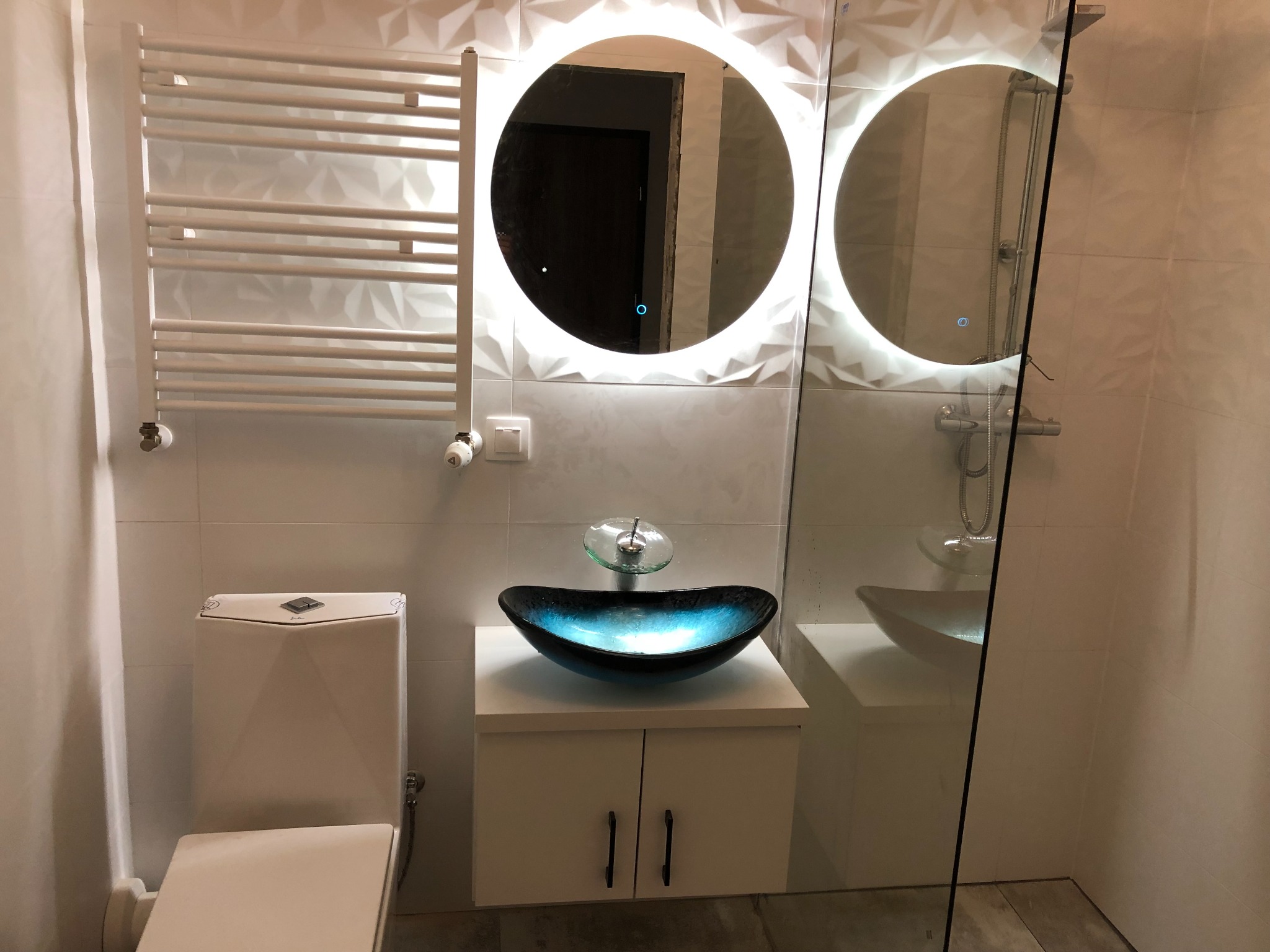 Nowoczesna łazienka z umywalką w kształcie misy w odcieniach błękitu i czerni, podświetlanym okrągłym lustrem i grzejnikiem drabinkowym na ścianie z trójwymiarową fakturą.