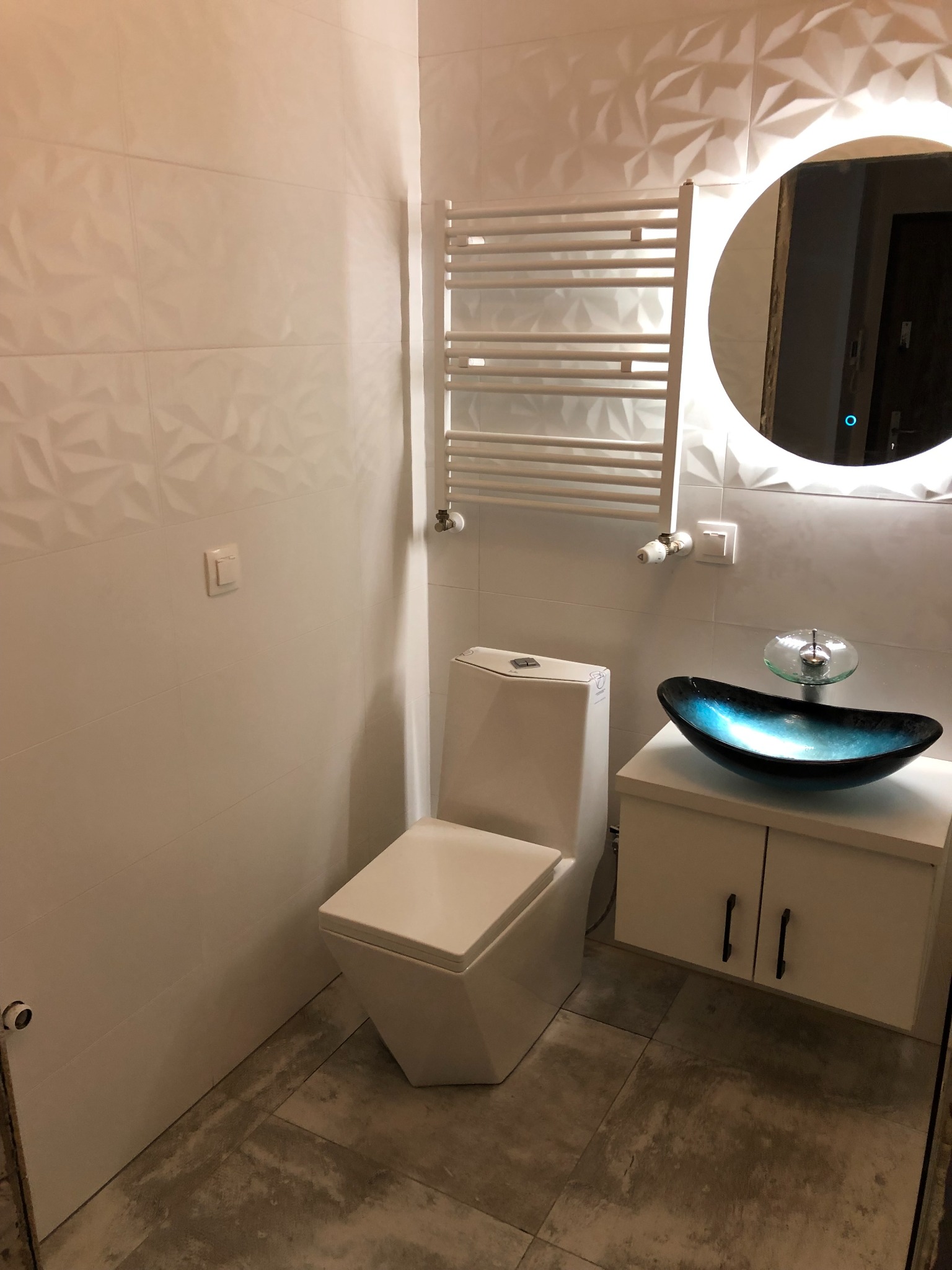 Nowoczesna łazienka z białymi, trójwymiarowymi płytkami, podświetlanym okrągłym lustrem, grzejnikiem drabinkowym, toaletą oraz umywalką w kształcie łodzi z niebieskiego szkła na białej szafce.