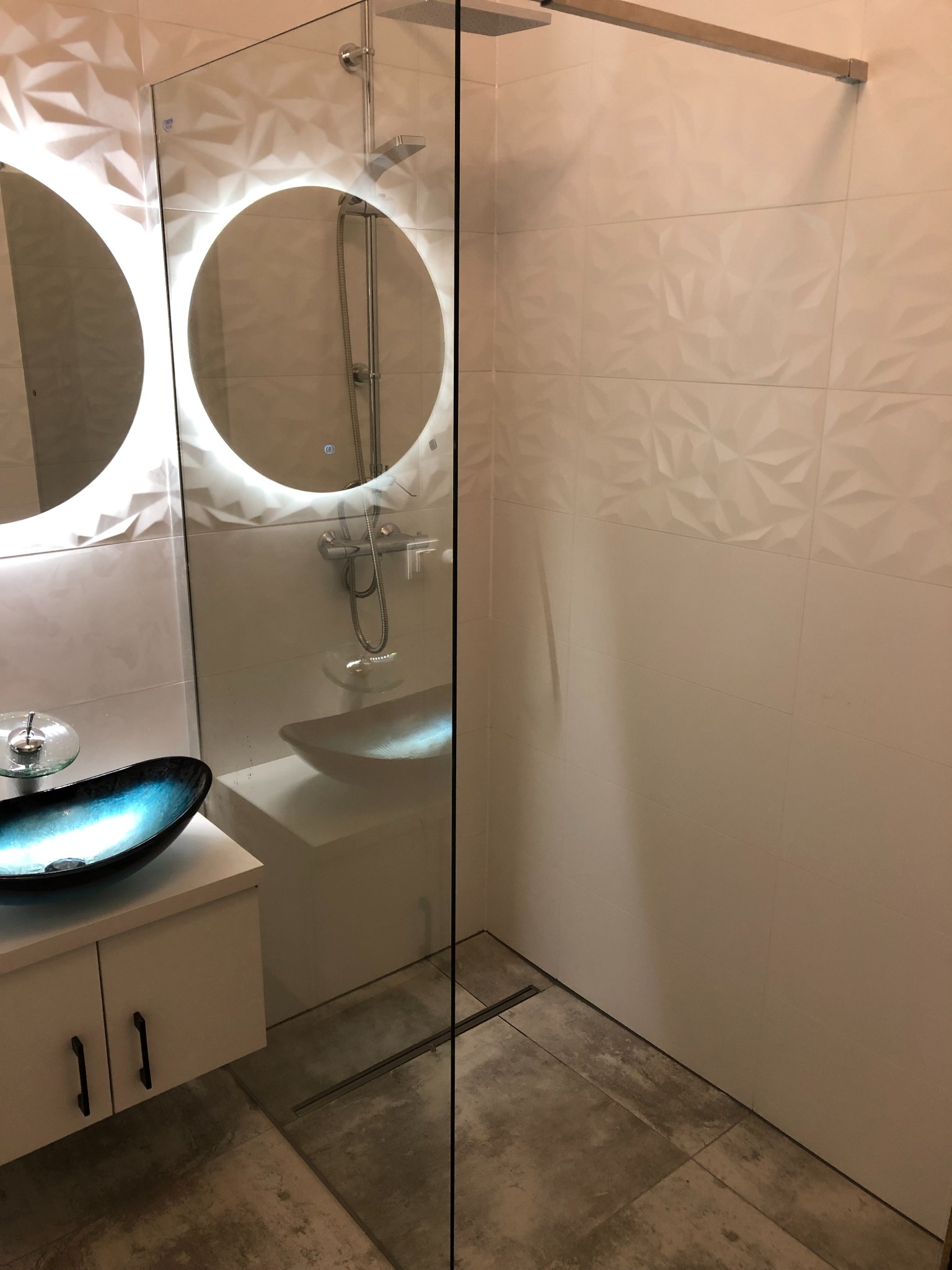 Nowoczesna łazienka z kabiną prysznicową typu walk-in, umywalką w kolorze turkusowym i okrągłymi lustrami z podświetleniem LED, wykończona białymi płytkami 3D.