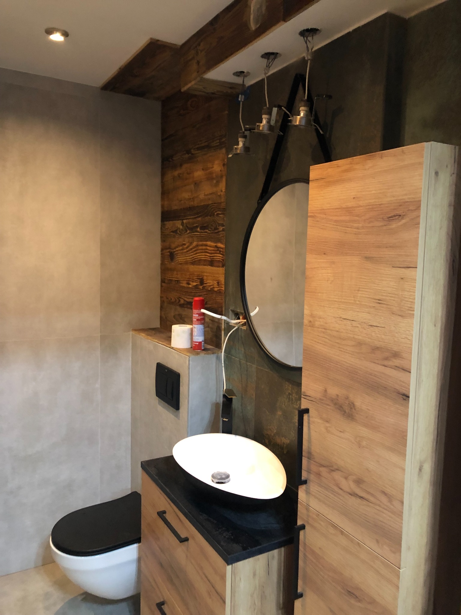 Nowoczesna łazienka z elementami drewna: wisząca toaleta z czarną deską, owalna umywalka na czarnym blacie, szafka z drewnianymi frontami, okrągłe lustro i surowa ściana z drewnianymi belkami.