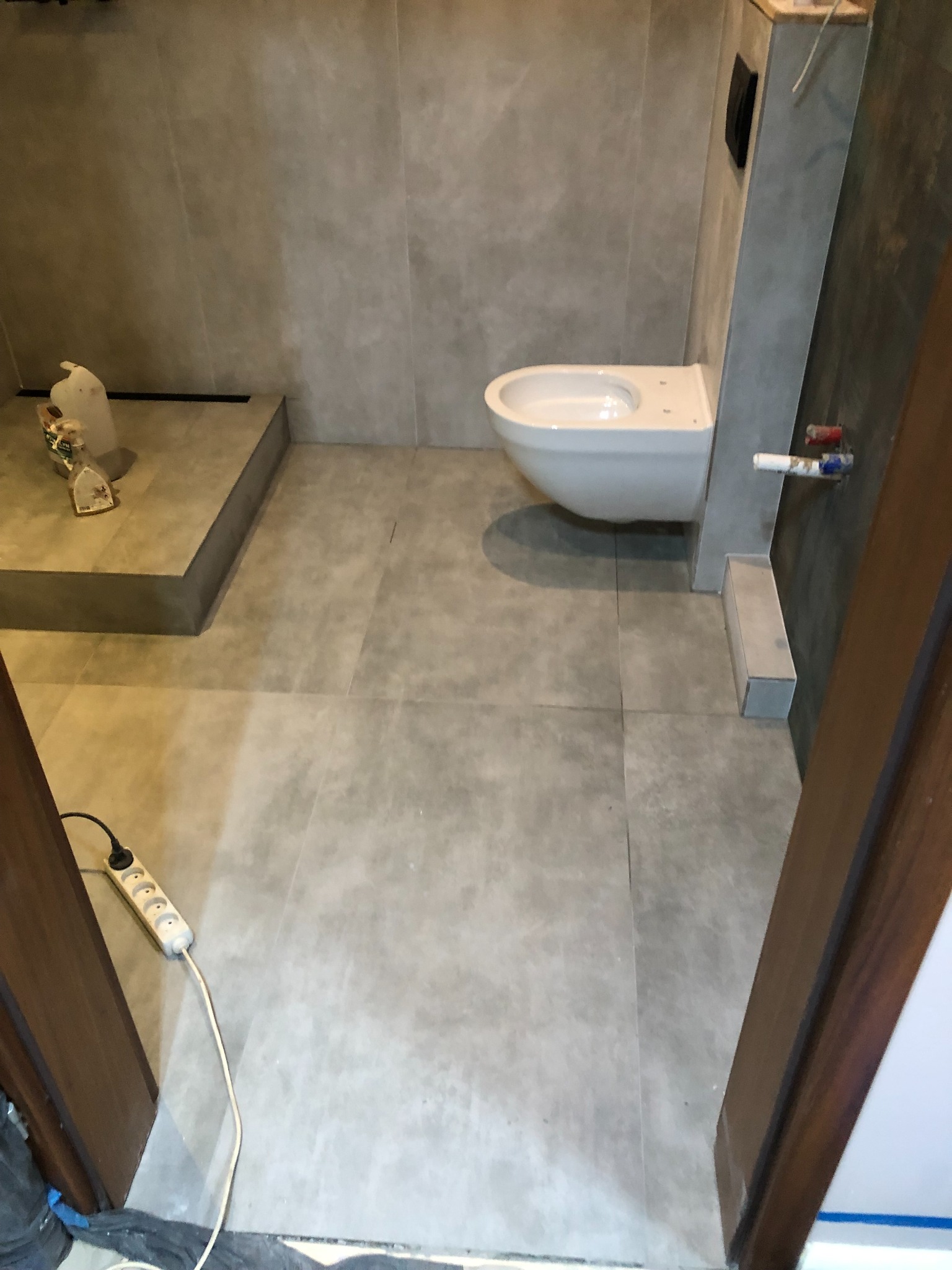 Łazienka w trakcie prac wykończeniowych, widoczna podwieszana toaleta, szare płytki na ścianach i podłodze, listwa przypodłogowa, przedłużacz na podłodze, w tle wiadro z zaprawą.
