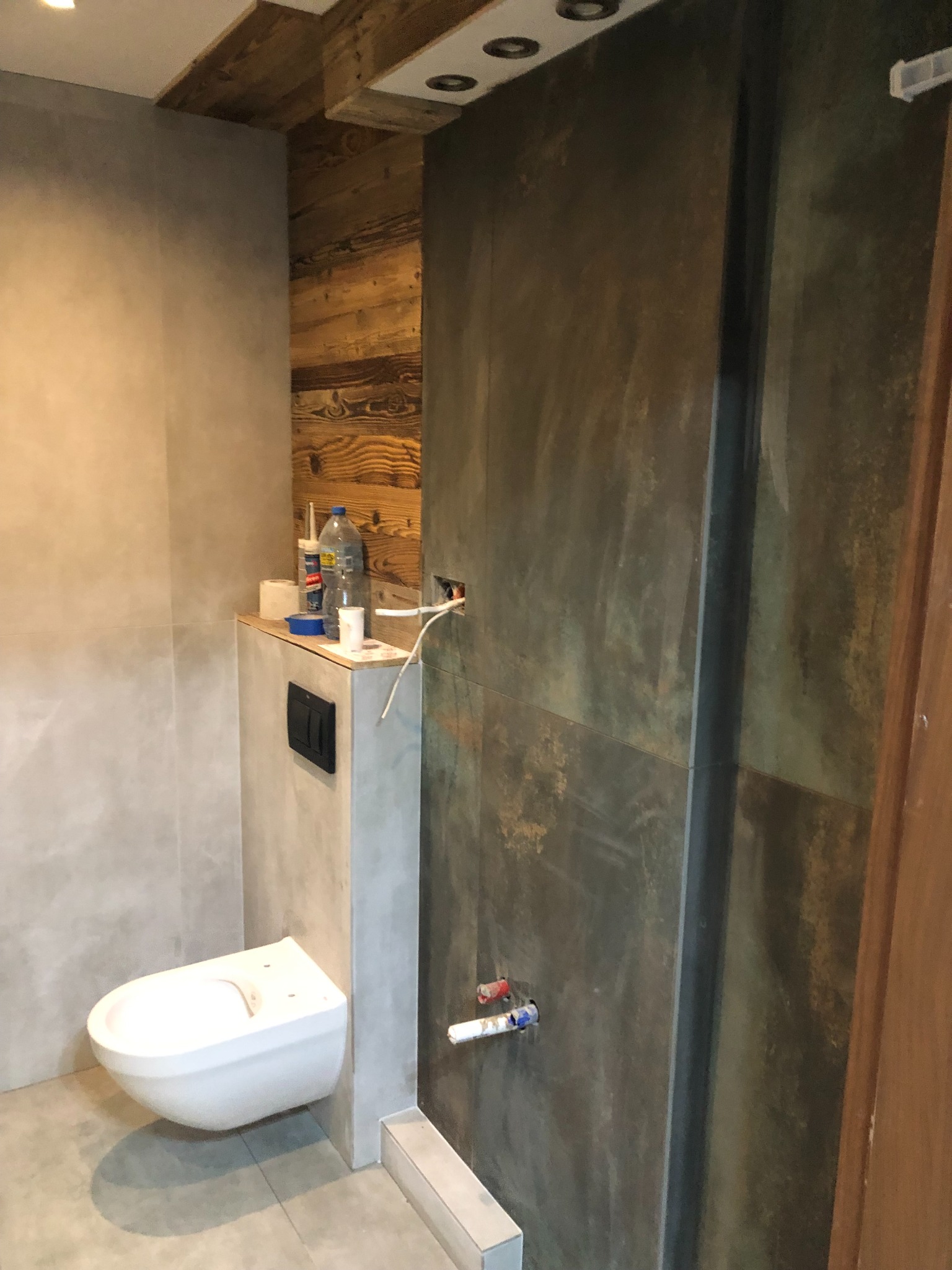 Wnętrze łazienki w trakcie prac wykończeniowych: podwieszana toaleta, ściana z ciemnych płytek imitujących rdzę, drewniana boazeria pod sufitem, widoczne rury i kable przed podłączeniem.