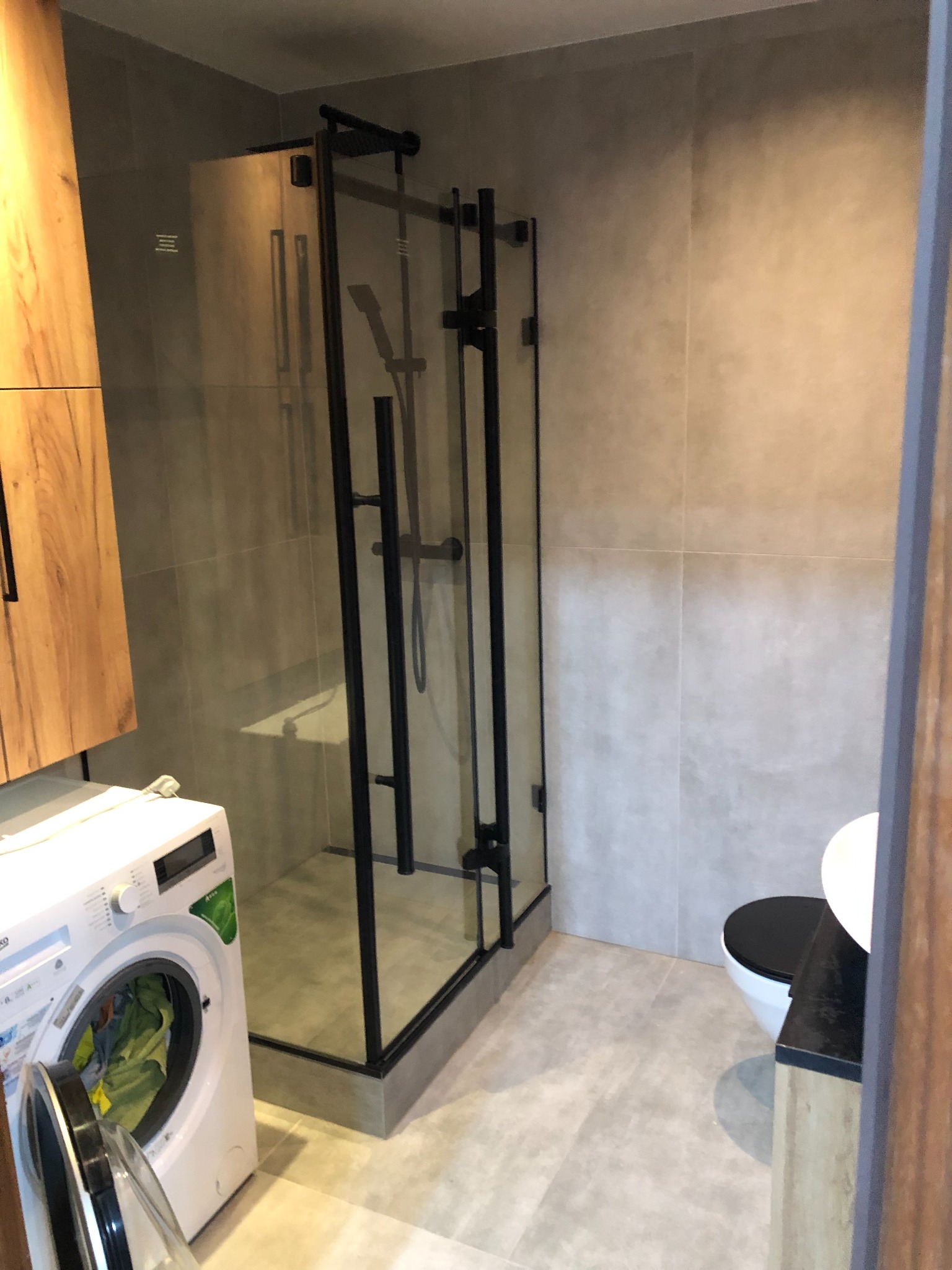 Nowoczesna łazienka z kabiną prysznicową typu walk-in z czarnymi ramami, pralką Beko i wiszącą toaletą z czarną deską, wykończona płytkami w odcieniach szarości.