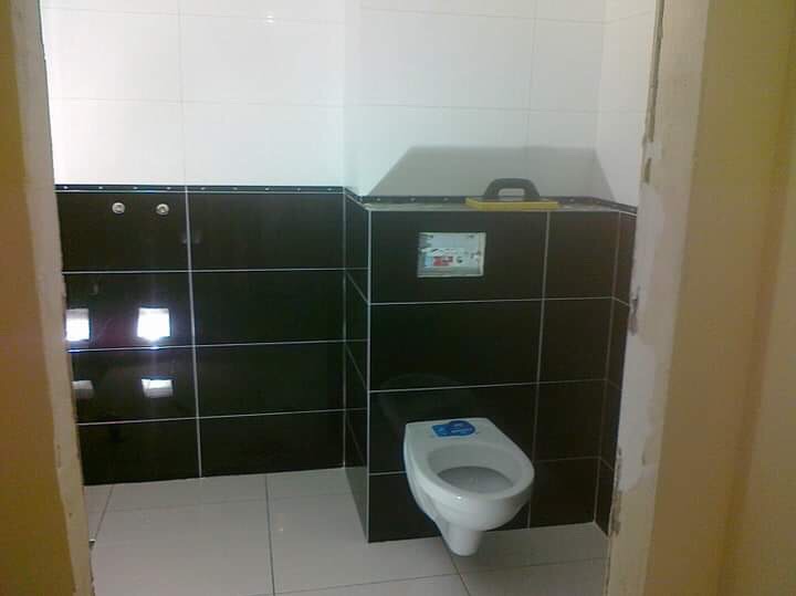 Częściowo wykończona łazienka z czarnymi i białymi płytkami, wisząca toaleta z niebieską naklejką na desce, żółta paca na górnej krawędzi płytek.