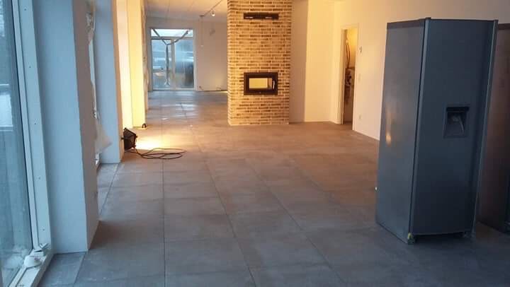 Nowa szara podłoga z płytek w przestronnym, jasnym pomieszczeniu z ceglanym kominkiem i dużymi oknami, widoczne przygotowania do dalszych prac wykończeniowych.