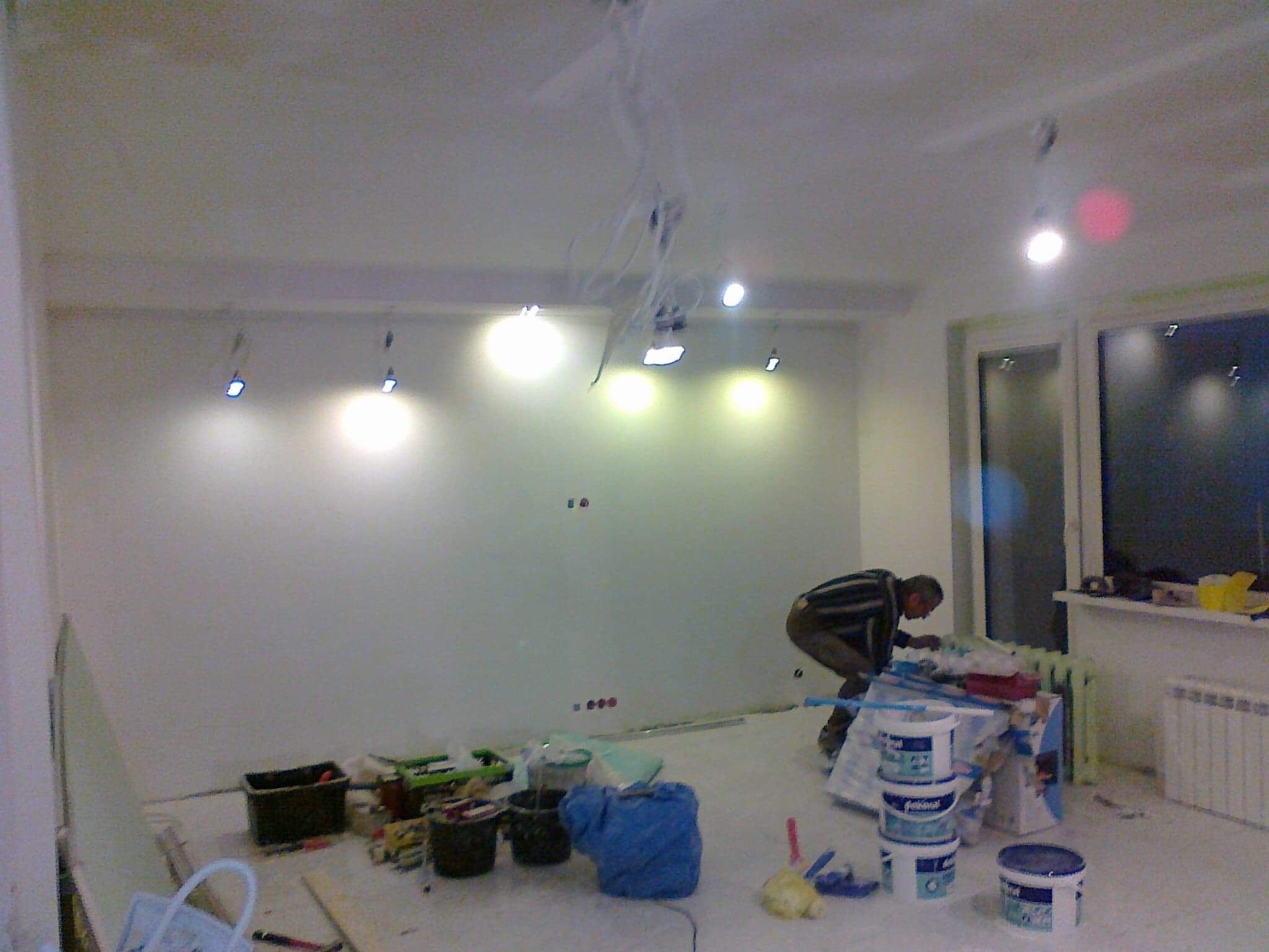 Pokój w trakcie przygotowań do malowania: widoczne puszki z farbą, narzędzia malarskie i instalacja oświetleniowa na suficie z wiszącymi kablami, mężczyzna pochylony nad pracami przygotowawczymi.