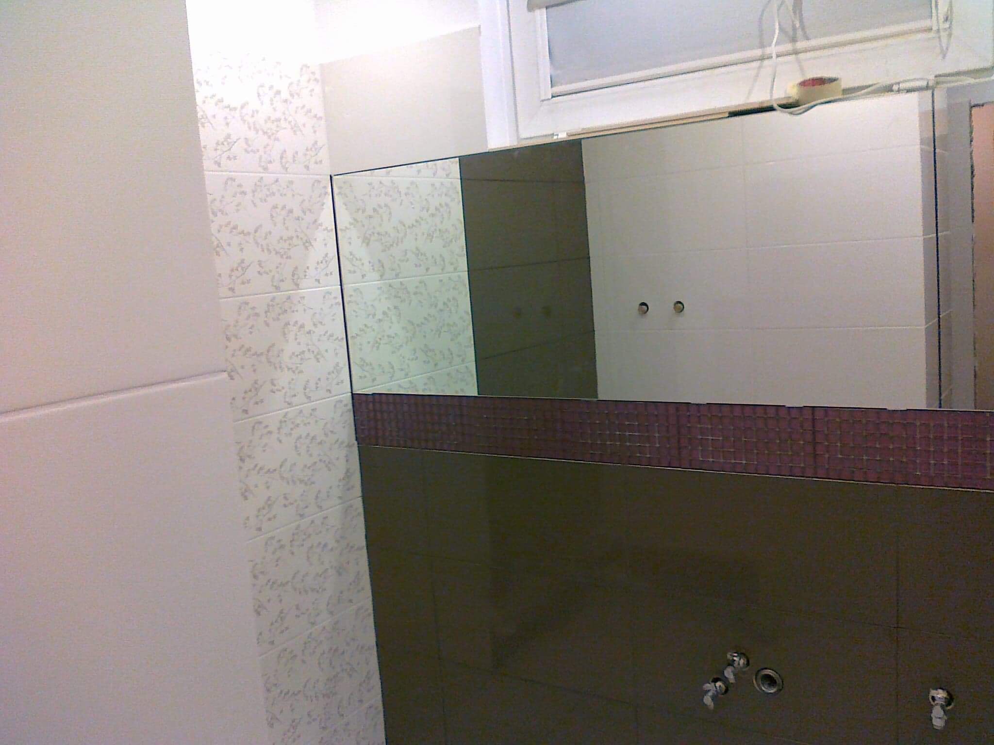 Fragment wykończonej łazienki z lustrem, widoczne płytki ścienne z motywem roślinnym, szare płytki oraz bordowa mozaika, bateria umywalkowa i odpływ podłogowy.