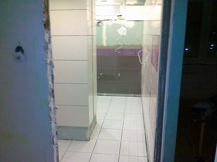 Wnętrze łazienki w trakcie remontu: białe płytki na ścianach i podłodze, widoczne ślady po szpachlowaniu na ścianie, fragment instalacji elektrycznej, surowe wykończenie.