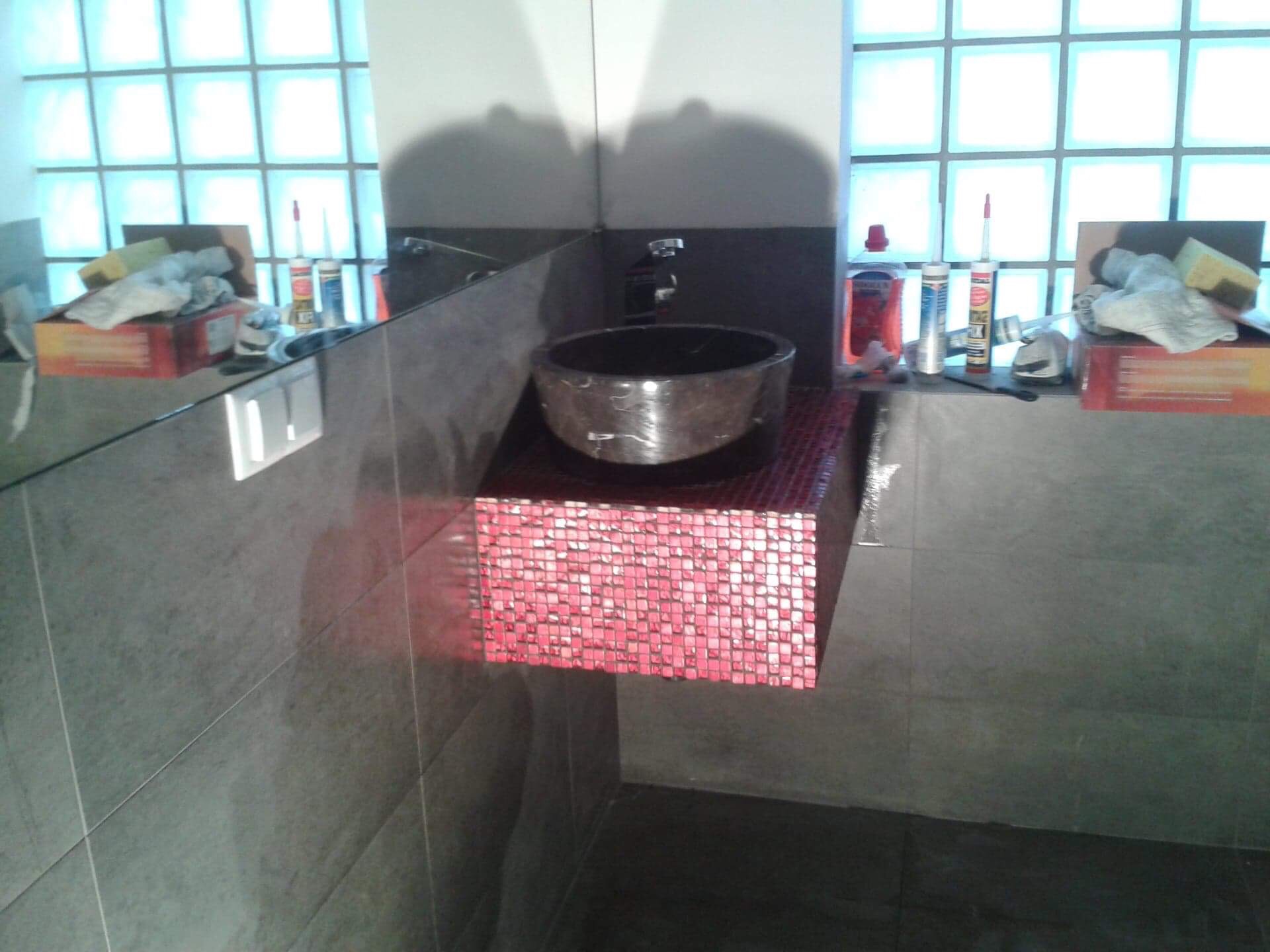 Umywalka nablatowa z ciemnego kamienia, osadzona na blacie wykończonym czerwoną mozaiką w łazience w Bydgoszczy, widoczne materiały budowlane i okno z luksferów.