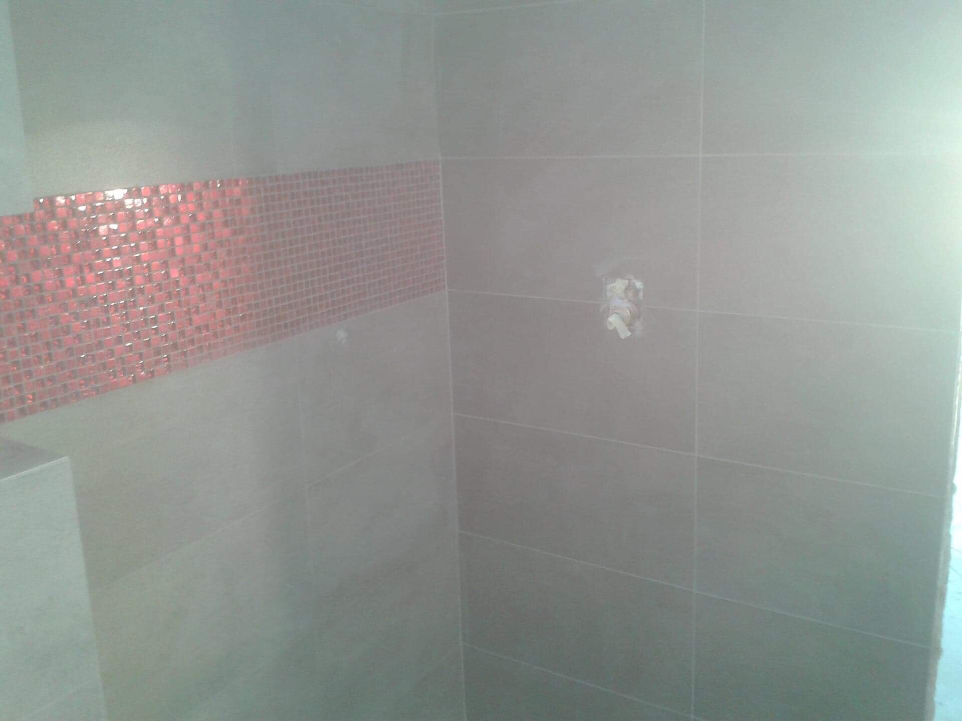 Ściana łazienki wyłożona szarymi płytkami ceramicznymi z poziomym pasem dekoracyjnej, czerwonej mozaiki szklanej, widoczny otwór na rurę.