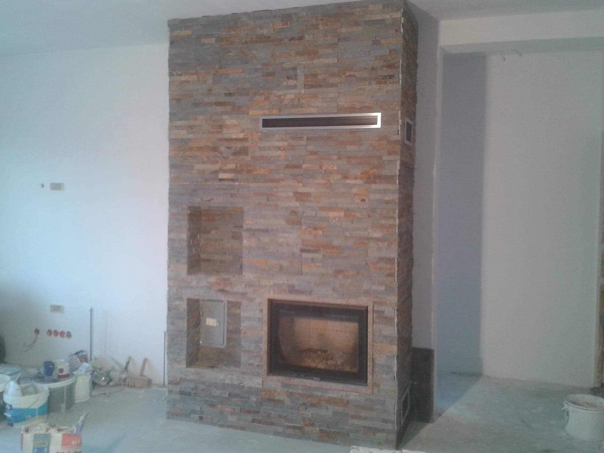 Obudowa kominka z kamienia dekoracyjnego w trakcie montażu, widoczne otwory wentylacyjne i puste wnęki, w tle ściany w trakcie malowania i narzędzia pracy.