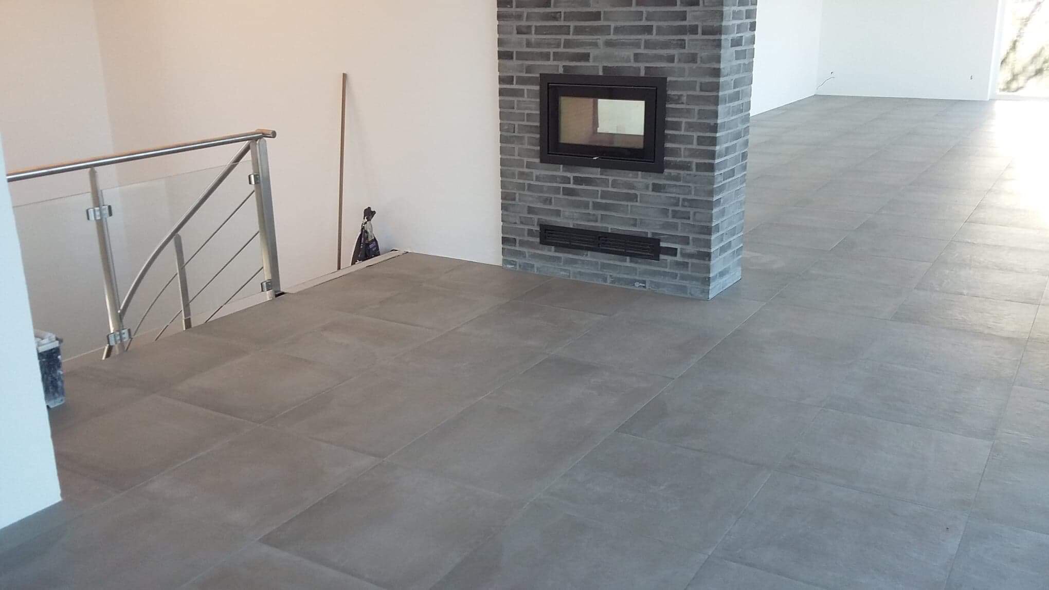 Wyłożona szarymi płytkami podłoga z kominkiem obudowanym szarą cegłą i szklaną balustradą schodów w jasnym wnętrzu.